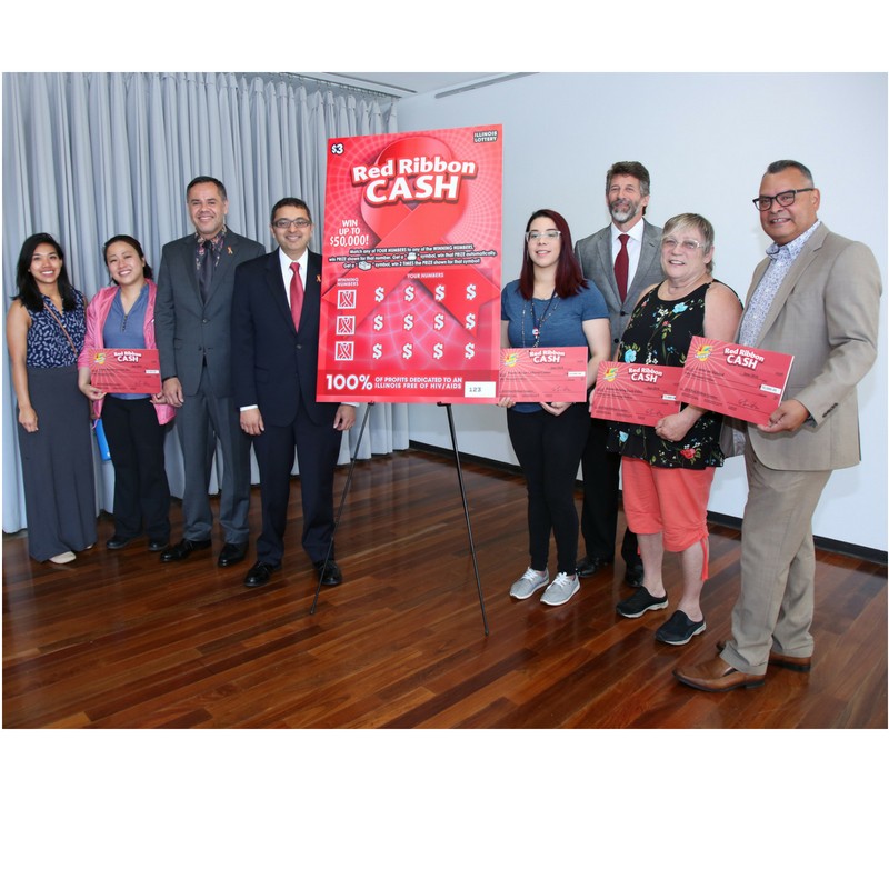 Greg Smith, Acting Director, Illinois Lottery, Modesto Valle, CEO, Center on Halsted, Illinois Department of Public Health (IDPH) Director Nirav D. Shah, M.D., J.D., and Eduardo Alvarado, HIV/AIDS Section, IDPH stand with the newly unveiled 2018 Red Ribbon Cash instant ticket and representatives from four out of 11 the 2018 grant recipients.